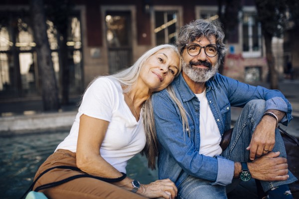 A happy mature couple sits together outdoors. The woman, with long white hair, leans on the man's shoulder. The man has gray curly hair and a beard, wearing glasses and a denim shirt.