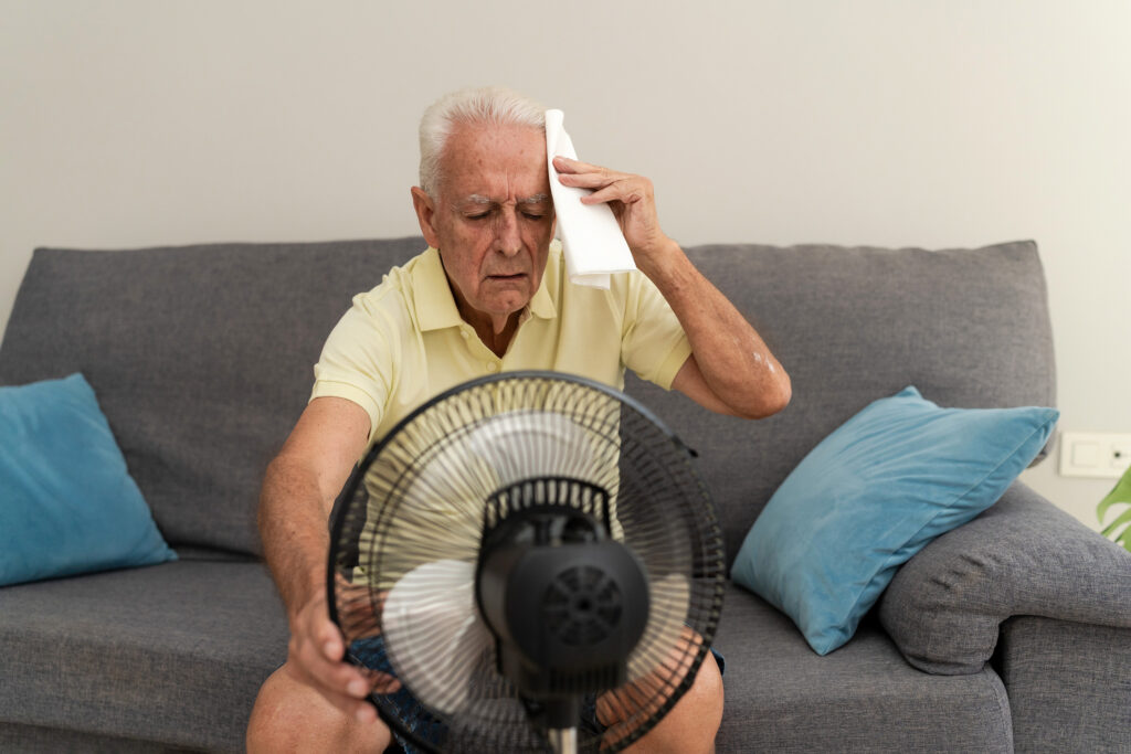 Elderly man cooling off with electric fan and wiping sweat from forehead suffering from summer heatstroke